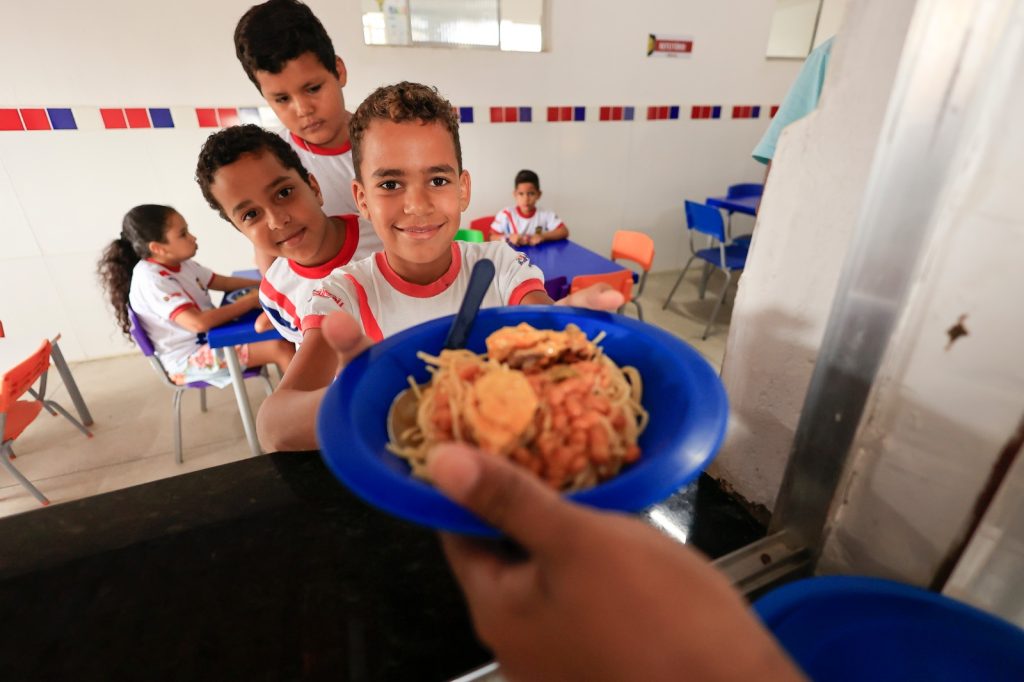 Bom dia, Inácio! 😊 Aqui está uma descrição acessível da imagem: Um grupo de crianças está em um refeitório escolar. Três delas estão na fila do balcão de comida, e uma está recebendo um prato azul com espaguete, feijão e um pedaço de ovo cozido. Todas usam uniforme branco com detalhes vermelhos e azuis. Ao fundo, outras crianças estão sentadas em cadeiras coloridas, comendo ou esperando a refeição. As paredes têm azulejos nas cores vermelho, branco e azul, e há uma placa com a palavra “REFEITÓRIO”. Se quiser, posso adaptar essa descrição para diferentes públicos ou formatos!