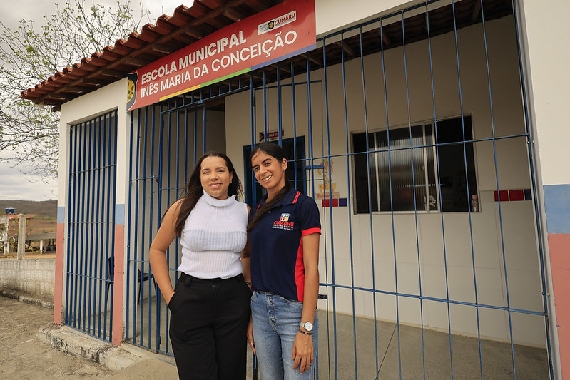 A foto mostra Amanda Joelly e Nadjane de Moura em frente à Escola Municipal Inês Maria da Conceição. Amanda, à esquerda, veste uma blusa branca e sorri suavemente. Nadjane, à direita, usa uma camiseta azul escura e também está sorridente. As duas estão atrás de um portão azul, com a fachada da escola ao fundo — pintada em branco, azul e rosa, com telhado de cerâmica. A placa da escola está visível acima delas, destacando o nome da instituição e o logotipo da prefeitura.