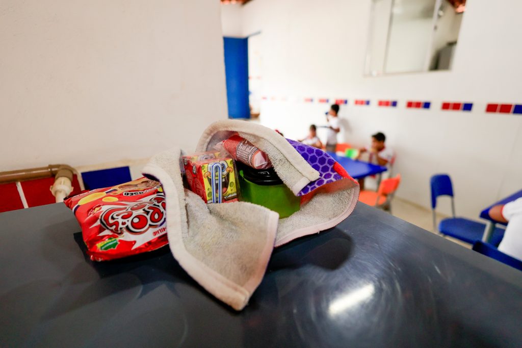 A foto mostra um pacote de lanche infantil disposto sobre uma superfície escura, envolto em uma toalha. Dentro do pacote, há um saquinho vermelho de balas de goma da marca, uma caixinha de suco decorada com personagens de desenho animado coloridos, um recipiente verde com tampa preta e outros itens de lanche não totalmente visíveis. Ao fundo, vê-se um ambiente claro com paredes brancas e detalhes em azulejos vermelhos e azuis. Crianças estão sentadas em mesas com cadeiras coloridas — azul, laranja e vermelha — sugerindo que o local é uma creche ou escola, possivelmente durante o horário do lanche.