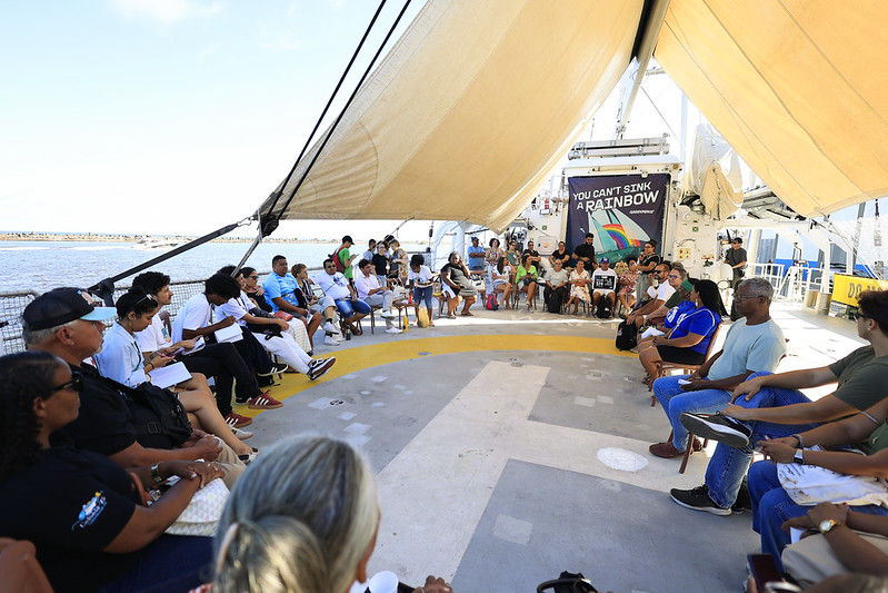 A imagem mostra um grupo grande de pessoas sentadas em círculo sobre o convés de um navio, sob uma cobertura ampla que protege do sol. A reunião parece ser um encontro, roda de conversa ou oficina, com clima colaborativo e informal. Ao fundo, vê-se o mar e uma faixa colorida com a frase “YOU CAN’T SINK A RAINBOW” (em português: “Você não pode afundar um arco-íris”), acompanhada de um desenho de arco-íris e o nome “GREENPEACE”, indicando que o evento está ligado à organização ambiental. O convés tem uma marcação circular amarela com uma seta, sugerindo que também serve como heliponto