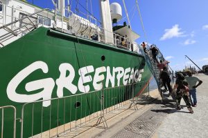 A imagem mostra um grande navio verde atracado em um porto. Na lateral do navio, está pintada em letras brancas a palavra GREENPEACE, indicando que pertence à organização ambiental de mesmo nome. O dia está ensolarado, com céu azul claro. Algumas pessoas estão subindo por uma passarela para embarcar no navio, enquanto outras permanecem no cais, observando ou aguardando. O navio possui equipamentos marítimos visíveis, como botes salva-vidas e antenas de comunicação.