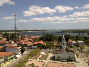 A foto mostra uma vista panorâmica de Jericoacoara, uma charmosa vila litorânea. No centro, destaca-se uma igreja com torre alta, cercada por casas de telhados vermelhos. Ao fundo, vê-se uma grande extensão de água — provavelmente o mar ou uma lagoa — que emoldura a paisagem. A vegetação é abundante, com muitas árvores e palmeiras espalhadas pela vila. À esquerda, há uma torre de telecomunicações, sinalizando a presença de infraestrutura moderna. O céu está claro, com algumas nuvens, indicando um dia ensolarado e tranquilo.