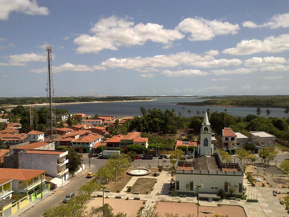 A foto mostra uma vista panorâmica de Jericoacoara, uma charmosa vila litorânea. No centro, destaca-se uma igreja com torre alta, cercada por casas de telhados vermelhos. Ao fundo, vê-se uma grande extensão de água — provavelmente o mar ou uma lagoa — que emoldura a paisagem. A vegetação é abundante, com muitas árvores e palmeiras espalhadas pela vila. À esquerda, há uma torre de telecomunicações, sinalizando a presença de infraestrutura moderna. O céu está claro, com algumas nuvens, indicando um dia ensolarado e tranquilo.
