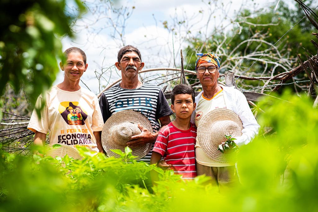 Ao ar livre, em um espaço verde com cerca de madeira ao fundo, três adultos - duas mulheres e um homem) e uma criança estão de pé, vestidos de forma casual. Um dos adultos segura folhas verdes, outro usa (ou segura) chapéu de palha, e há referência a “II Feira de Economia Solidária” em uma camiseta.