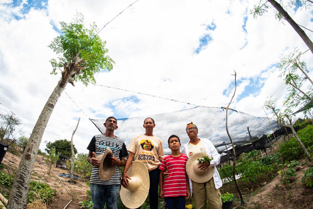 Quatro pessoas estão em pé em uma área de cultivo ao ar livre, sob um céu parcialmente nublado. São três adultos e uma criança, todos segurando chapéus de palha. Um dos adultos também segura um pequeno ramo de plantas verdes. A pessoa ao centro veste uma camiseta com os dizeres “Feira da Economia Solidária”, indicando participação em uma atividade comunitária ligada à agricultura sustentável. Ao fundo, há plantas cultivadas e uma estrutura com tela, possivelmente usada para proteção ou sombreamento.