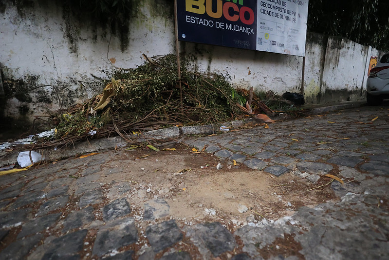 A imagem mostra uma rua de paralelepípedos com um trecho danificado, onde faltam algumas pedras e há terra exposta. Ao lado da rua, vê-se um monte de galhos, folhas e restos de poda, indicando descarte de resíduos orgânicos. Atrás do monte, há um muro branco com manchas e sinais de desgaste. Acima, um grande cartaz está fixado em estruturas metálicas. O cartaz traz o título PERNAMBUCO – Estado de Mudança e menciona um projeto relacionado à cidade de Olinda, PE, envolvendo geocompostagem e gestão urbana. À direita, aparece parcialmente um carro branco estacionado na calçada.