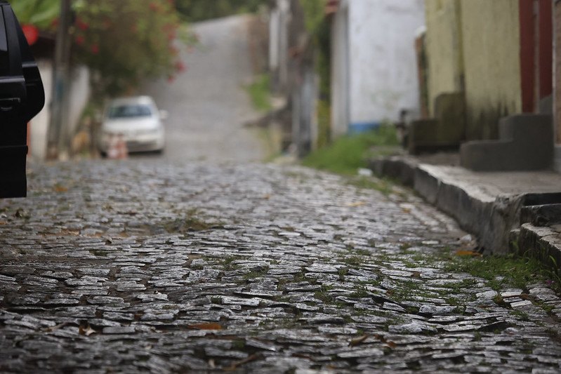 A imagem mostra uma rua de paralelepípedos em uma área residencial, vista de um ângulo baixo próximo ao chão. Os paralelepípedos estão molhados, indicando chuva recente, e há pequenos tufos de grama entre eles. À esquerda, parte de um carro preto é visível; mais adiante, um carro branco está estacionado próximo a cones laranja. A rua tem uma leve inclinação e é ladeada por casas com fachadas coloridas. O fundo desfocado destaca o relevo e a textura das pedras no primeiro plano.