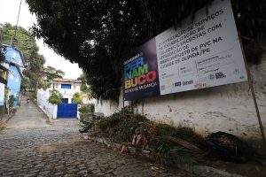 A imagem mostra uma rua de paralelepípedos com um trecho danificado, onde faltam algumas pedras e há terra exposta. Ao lado da rua, vê-se um monte de galhos, folhas e restos de poda, indicando descarte de resíduos orgânicos. Atrás do monte, há um muro branco com manchas e sinais de desgaste. Acima, um grande cartaz está fixado em estruturas metálicas. O cartaz traz o título PERNAMBUCO – Estado de Mudança e menciona um projeto relacionado à cidade de Olinda, PE, envolvendo geocompostagem e gestão urbana. À direita, aparece parcialmente um carro branco estacionado na calçada.