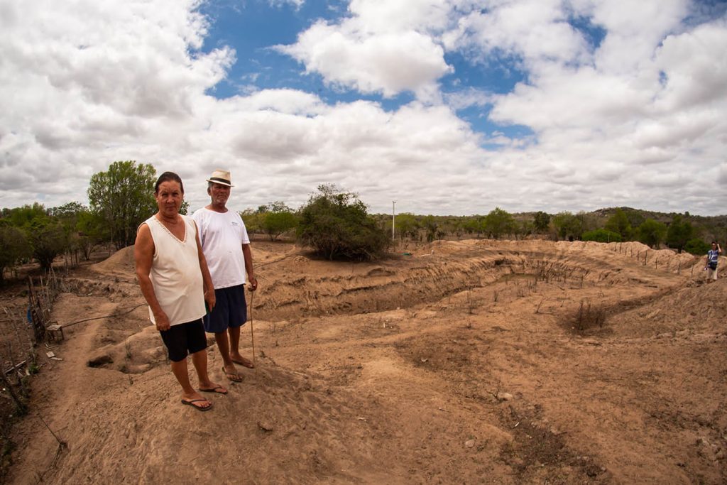 Duas pessoas - uma mulher e um homem - estão em pé sobre um terreno seco e irregular, com sulcos profundos e pouca vegetação. O solo parece árido, com algumas árvores verdes ao fundo e o céu parcialmente nublado. Ambas estão vestidas de forma simples, apropriadas para clima quente, e uma delas segura um bastão de caminhada. A paisagem sugere uma área rural afetada por erosão ou seca.
