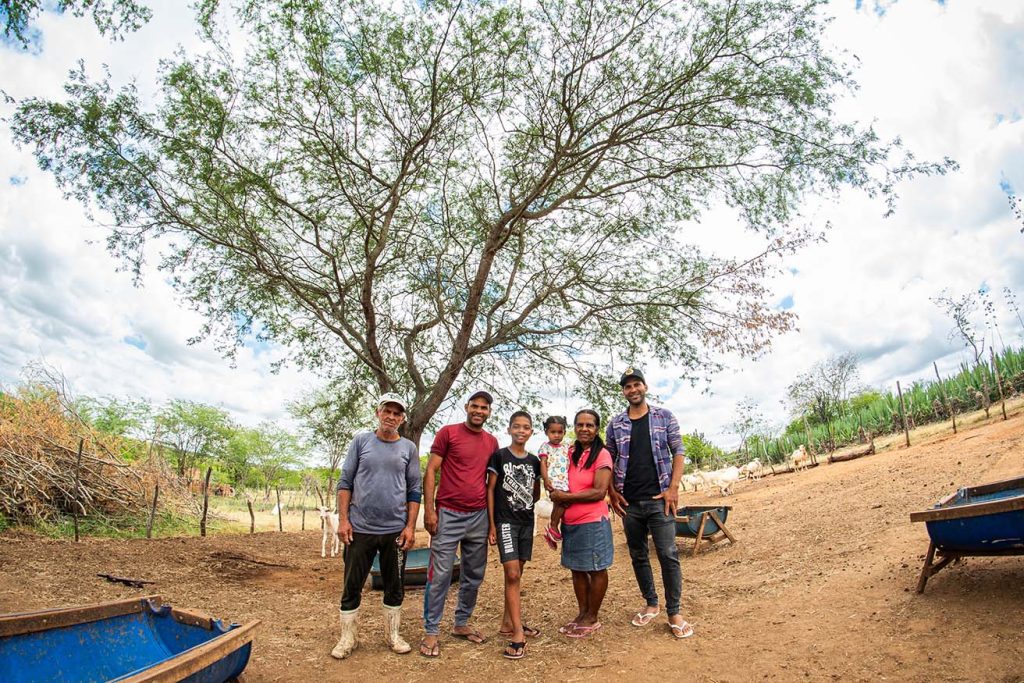 Família de agricultores, pai, mãe, dois filhos e dois netos, pousa para foto em um curral de cabras. Eles estão embaixo de um árvore.
