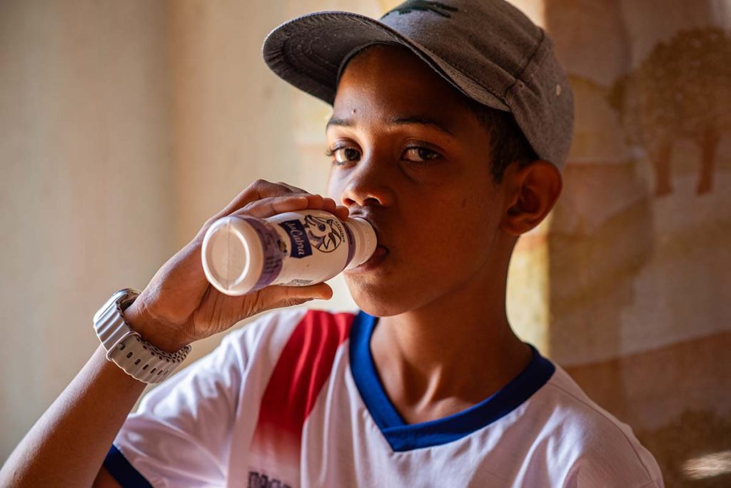 Menino de 10 anos, usando boné e fardamento escolar, toma uma garrafinha de iogurte de leite de cabra
