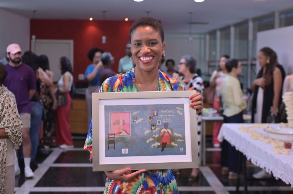 A escritora Luana Tolentino aparece sorridente em um ambiente iluminado, segurando um quadro com a ilustração da capa do livro Mamãe Aprendeu a Ler. Ela está em pé, vestida de forma elegante e parece estar participando de um evento especial. Ao fundo, há pessoas conversando e uma mesa com comidas e bebidas, sugerindo um clima de celebração.