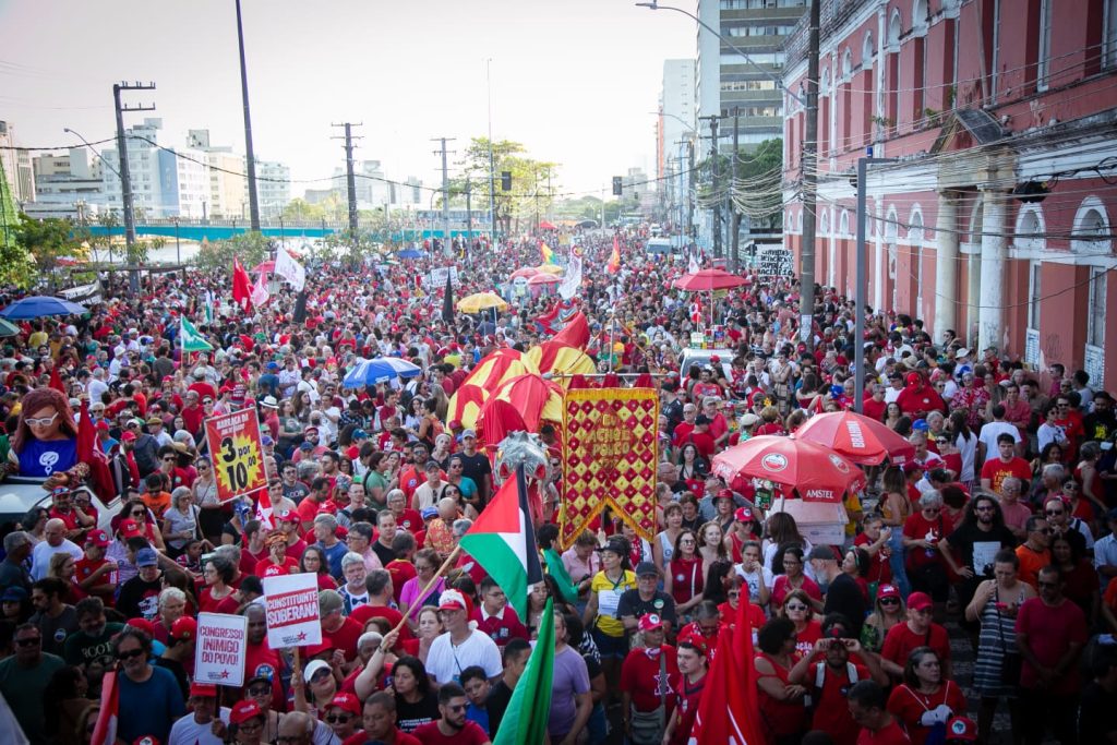 A imagem mostra uma grande manifestação popular em uma rua urbana. Há uma multidão de pessoas vestidas principalmente de vermelho, segurando faixas, cartazes e bandeiras. Algumas mensagens visíveis incluem “Congresso inimigo do povo” e “Constituinte soberana”. O ambiente é vibrante e colorido, com guarda-chuvas decorativos e elementos festivos. Ao fundo, há prédios altos e uma construção colonial vermelha à direita. A cena transmite um forte senso de mobilização social e expressão política coletiva.