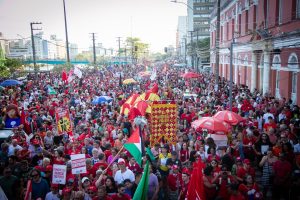 A imagem mostra uma grande manifestação popular em uma rua urbana. Há uma multidão de pessoas vestidas principalmente de vermelho, segurando faixas, cartazes e bandeiras. Algumas mensagens visíveis incluem “Congresso inimigo do povo” e “Constituinte soberana”. O ambiente é vibrante e colorido, com guarda-chuvas decorativos e elementos festivos. Ao fundo, há prédios altos e uma construção colonial vermelha à direita. A cena transmite um forte senso de mobilização social e expressão política coletiva.