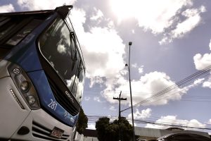 Um ônibus azul e branco está parado sob um céu com nuvens iluminadas pelo sol. Ao fundo, há postes, árvores e um prédio urbano compondo o cenário.
