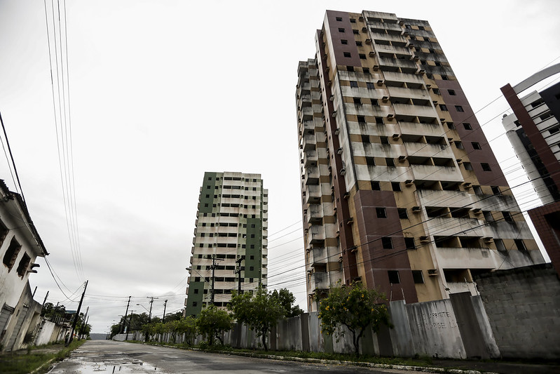 A imagem mostra dois prédios altos e antigos, com vários andares e sacadas, que parecem estar abandonados ou mal conservados. Um deles tem fachada marrom e bege; o outro, verde e bege. Muitas janelas estão escuras ou quebradas, e as sacadas estão vazias. A rua em frente está molhada, como se tivesse chovido, e não há pessoas nem carros. Há árvores pequenas ao lado de um muro de concreto e fios de energia atravessam a cena. O clima é de abandono e silêncio.