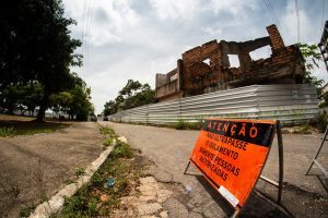 A foto mostra um prédio antigo de tijolos parcialmente destruído, cercado por uma barreira metálica para segurança. Na frente, há uma placa de alerta laranja com texto em português que diz: “Atenção: Não ultrapasse o isolamento. Somente pessoas autorizadas.” O local está ao lado de uma calçada pavimentada com árvores à esquerda e o céu está nublado ao fundo.