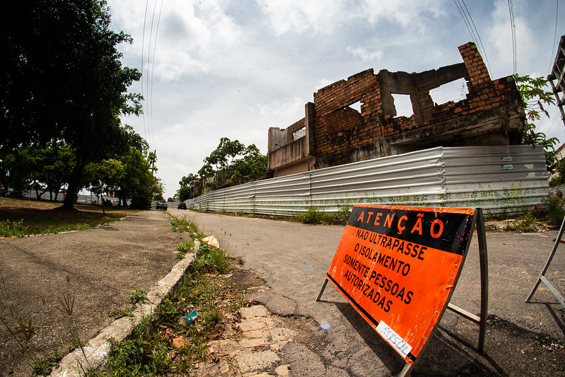 A foto mostra um prédio antigo de tijolos parcialmente destruído, cercado por uma barreira metálica para segurança. Na frente, há uma placa de alerta laranja com texto em português que diz: “Atenção: Não ultrapasse o isolamento. Somente pessoas autorizadas.” O local está ao lado de uma calçada pavimentada com árvores à esquerda e o céu está nublado ao fundo.