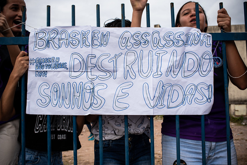 Um grupo de pessoas está reunido atrás de uma grade metálica azul, segurando um cartaz de protesto feito à mão. O cartaz é de papel branco com letras grandes e escuras escritas em português. Ele diz: BRASKEM ASSASSINA DESTRUINDO SONHOS E VIDAS! e #BRASKEM NÃO TOLERAREMOS!. A cena transmite indignação e resistência, com os manifestantes expressando sua oposição à empresa Braskem.