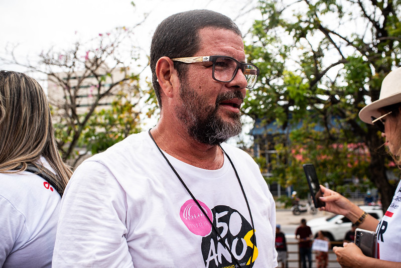 Descrição acessível: O pastor Wellington Santos aparece em destaque, usando óculos e barba, vestindo uma camiseta branca com a inscrição 50 ANOS e um adesivo rosa circular. Ele está ao ar livre, cercado por outras pessoas que também usam camisetas brancas. Algumas estão conversando ou tirando fotos. Ao fundo, há árvores, um prédio e carros estacionados, indicando um ambiente urbano.