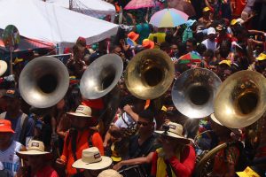 Uma multidão animada participa de um desfile de rua, com vários músicos tocando instrumentos de sopro (como tubas) em meio a cores vibrantes, chapéus e guarda-chuvas — clima de festa popular, tipo carnaval.