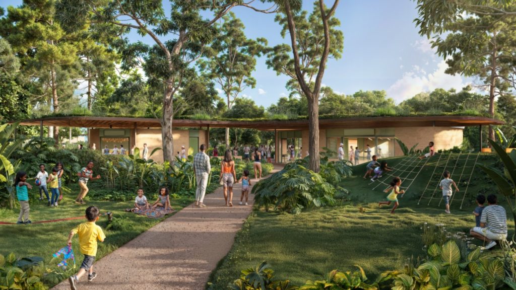 A imagem mostra uma cena alegre ao ar livre, provavelmente em um centro comunitário ou escola cercada por árvores altas e vegetação abundante. Ao fundo, há um prédio com telhado verde coberto por plantas, integrando-se ao ambiente natural. No gramado em frente, várias crianças brincam — algumas sobem em uma estrutura de cordas, outras correm ou participam de atividades diversas. Há também adultos caminhando, sentados ou interagindo entre si. O clima é de inclusão e convivência, com destaque para o contato com a natureza.