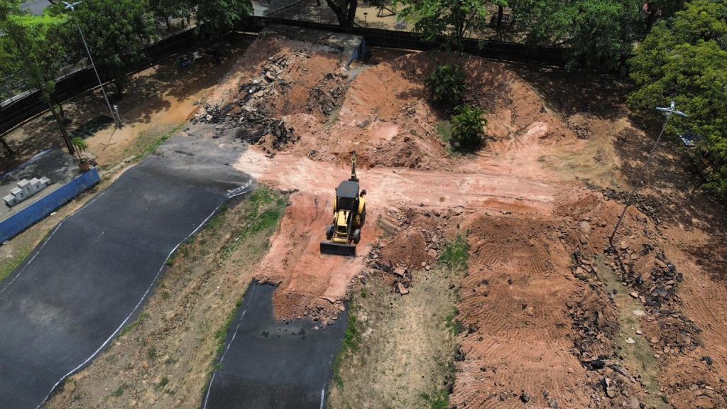 A foto mostra uma vista aérea de um canteiro de obras. No centro, há uma escavadeira amarela trabalhando sobre um caminho de terra avermelhada, movimentando o solo. Ao redor, vemos montes de terra e entulhos, indicando que a área está em processo de construção. De cada lado do caminho de terra, há partes de uma superfície pavimentada, possivelmente uma estrada, que parecem estar sendo modificadas. Árvores e uma cerca delimitam o espaço, sugerindo que o local está cercado e faz parte de um projeto maior.