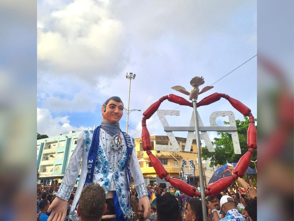 A imagem mostra um grande boneco humanoide sendo carregado em meio a uma multidão durante uma festa de rua. Ao lado dele há uma estrutura em forma de coração com elementos vermelhos e letras decorativas, enquanto pessoas ao redor celebram em um ambiente festivo ao ar livre.