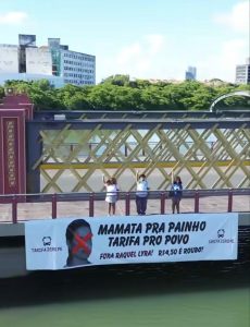 Três pessoas jovens estão em pé sobre uma ponte, atrás de uma faixa de protesto. A faixa tem uma foto de uma pessoa com um X vermelho sobre o rosto e frases escritas em português, criticando o valor da tarifa de transporte público e mencionando a governadora Raquel Lyra. Ao fundo, vê-se um céu claro, prédios e vegetação. A ponte passa sobre o rio Capibaribe.