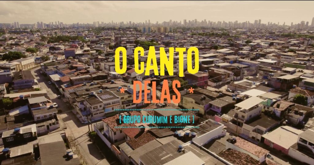 A imagem mostra uma vista aérea de uma área urbana densamente povoada, com muitas casas pequenas e prédios baixos de telhados planos, alguns com caixas d’água. Ao fundo, aparecem edifícios mais altos, indicando uma região central da cidade. Sobre a imagem, há um texto que diz: “O CANTO DELAS GRUPO CURUMIM E BIONE”.