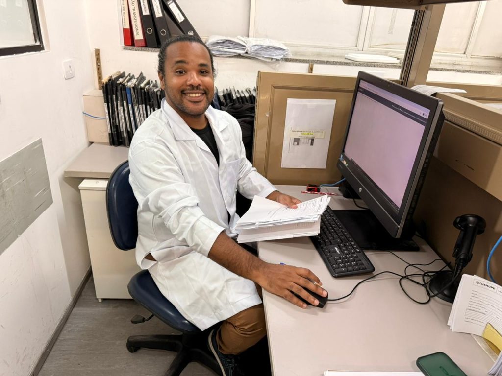 Imagem na horizontal de Paulo, um homem negro, sentado em uma cadeira de escritório, sorrindo para a câmera. Ele veste jaleco branco e está em frente a uma mesa com computador, teclado e mouse. Com a mão esquerda, segura uma pilha de papéis. Ao fundo, pastas e caixas organizadoras.