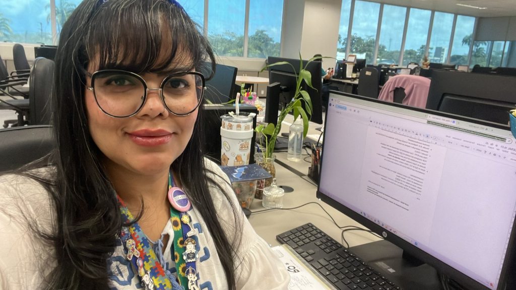 Imagem de Emmanuelle, uma mulher de pele clara, com cabelo liso preto e franja, usa óculos de armação redonda, um crachá com cordão colorido, broches e sorri levemente para a câmera. Ela está sentada de frente há um computador. Ao fundo, em um escritório amplo, com mesas, computadores e cadeiras ao fundo.