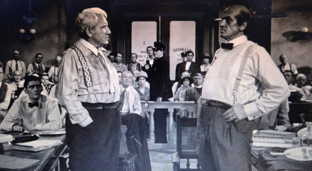 Na cena emblemática do tribunal em O Vento Será Tua Herança, dois advogados veteranos se encaram intensamente diante de uma sala lotada, simbolizando o confronto entre ciência e religião durante o famoso “Julgamento do Macaco”. Henry Drummond, defensor da liberdade de pensamento, encara Matthew Harrison Brady, representante da fé tradicional, em um momento de tensão dramática, com o público atento e a atmosfera carregada de expectativa, refletindo o embate ideológico que transcende o caso jurídico e ecoa questões universais sobre educação, crença e progresso.