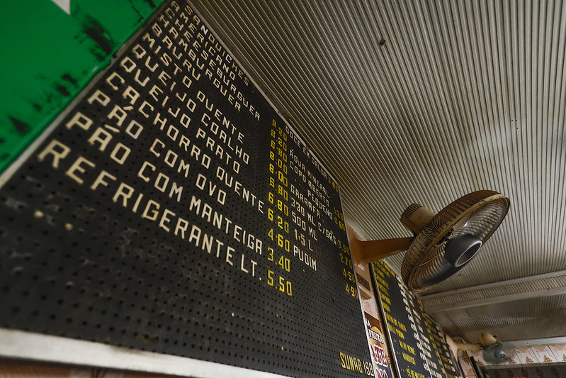 📋 A imagem mostra uma placa de cardápio fixada no teto de um estabelecimento simples, possivelmente uma lanchonete ou bar. A placa é preta com letras brancas e amarelas, organizadas em duas seções: “SANDUÍCHES” e “BEBIDAS”. Os itens incluem misto quente, cachorro-quente, pão com ovo, refrigerante, água com e sem gás, sucos, cervejas em diferentes tamanhos e garrafas de refrigerante. Os preços estão em reais e variam entre R$ 1,88 e R$ 13,00. Um ventilador de teto aparece ao lado da placa, reforçando o ambiente informal e funcional do local.