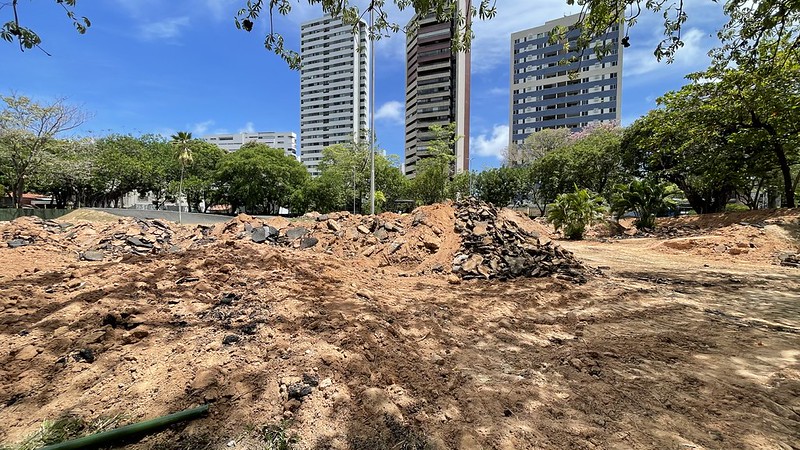 A foto mostra um local de escavação ou obra em uma área urbana. O chão está coberto por montes de terra e pedaços quebrados de asfalto ou concreto, indicando que o solo foi recentemente mexido. Ao fundo, vemos prédios altos cercados por árvores e vegetação, sugerindo que o espaço fica próximo a áreas residenciais ou comerciais. O céu está limpo com poucas nuvens, revelando um dia ensolarado.