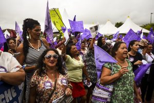 Uma multidão de mulheres participa de uma manifestação pública, segurando bandeiras roxas e panfletos. O ambiente é vibrante e engajado, com muitas delas usando adesivos e símbolos de um movimento coletivo. Ao fundo, há tendas brancas e outras bandeiras, reforçando o clima de mobilização social.