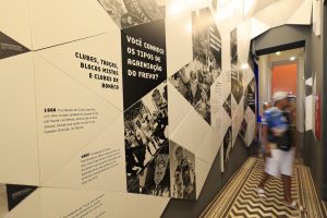 A foto mostra um corredor de exposição em um museu dedicado ao Frevo. Nas paredes, há painéis com textos e fotos históricas sobre clubes e agremiações carnavalescas. O espaço é decorado com padrões geométricos em preto e branco, criando contraste visual. O chão tem um desenho em zigue-zague que acompanha o estilo vibrante da mostra. Alguns visitantes caminham pelo corredor, observando os conteúdos expostos.