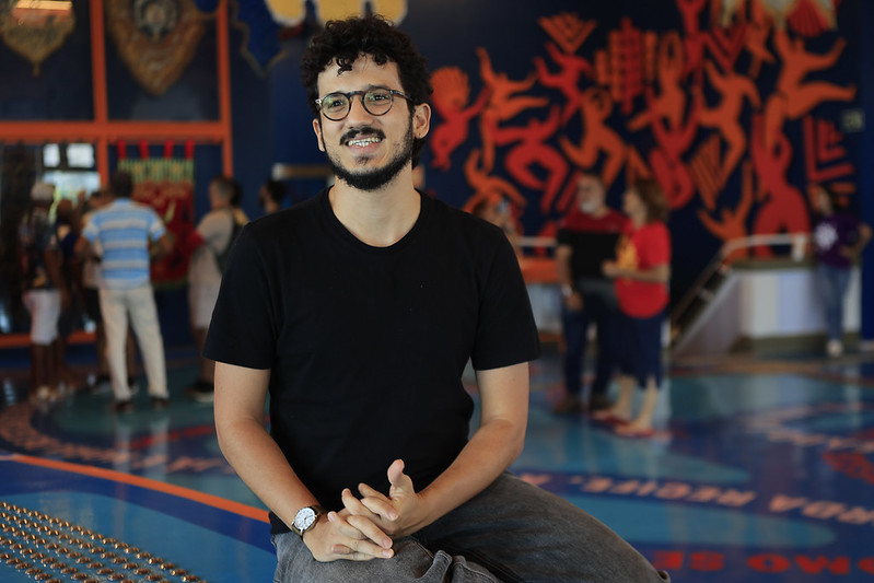 A foto mostra Luiz Vinicius Maciel, um homem de cabelos cacheados pretos, óculos redondo e barba, sentado e sorrindo em um espaço cultural colorido. Ele veste camiseta preta e calça cinza, transmitindo um ar descontraído. Ao fundo, há um mural vibrante em azul escuro com figuras vermelhas que remete a foliões de carnaval. Outras pessoas circulam pelo ambiente, que tem chão decorado com desenhos e textos, criando uma atmosfera artística e animada.