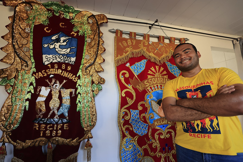 A imagem mostra Thomás Ricardo, um homem em pé, de braços cruzados, diante de dois estandartes festivos e ornamentados. Ele veste uma camiseta amarela com desenhos que combinam com os símbolos dos estandartes, indicando ligação com o grupo representado. Os estandartes são ricamente decorados em cores vivas como dourado, azul e vermelho, trazendo inscrições que mencionam “Vassourinhas” e “Recife”, além de figuras e emblemas. O conjunto transmite orgulho cultural e celebração de tradições locais.