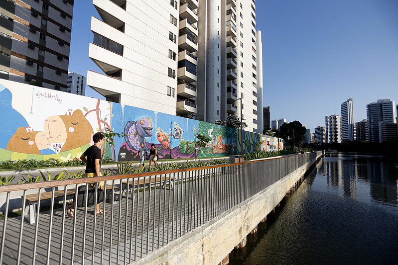 A imagem mostra uma área urbana moderna no Recife. Há prédios residenciais altos ao fundo, de aparência recente e cores neutras. Ao lado deles corre o rio Capibaribe, acompanhado por uma calçada para pedestres com grades metálicas de proteção. No primeiro plano, algumas pessoas caminham tranquilamente por essa via, aproveitando o espaço público. À esquerda, um longo muro está coberto por um mural colorido, cheio de desenhos artísticos — figuras de animais e formas abstratas — que dão vida e energia ao ambiente. A presença da água, da vegetação nas margens e da arte urbana cria uma atmosfera agradável, sugerindo que o local foi planejado para ser mais acolhedor e culturalmente vibrante.