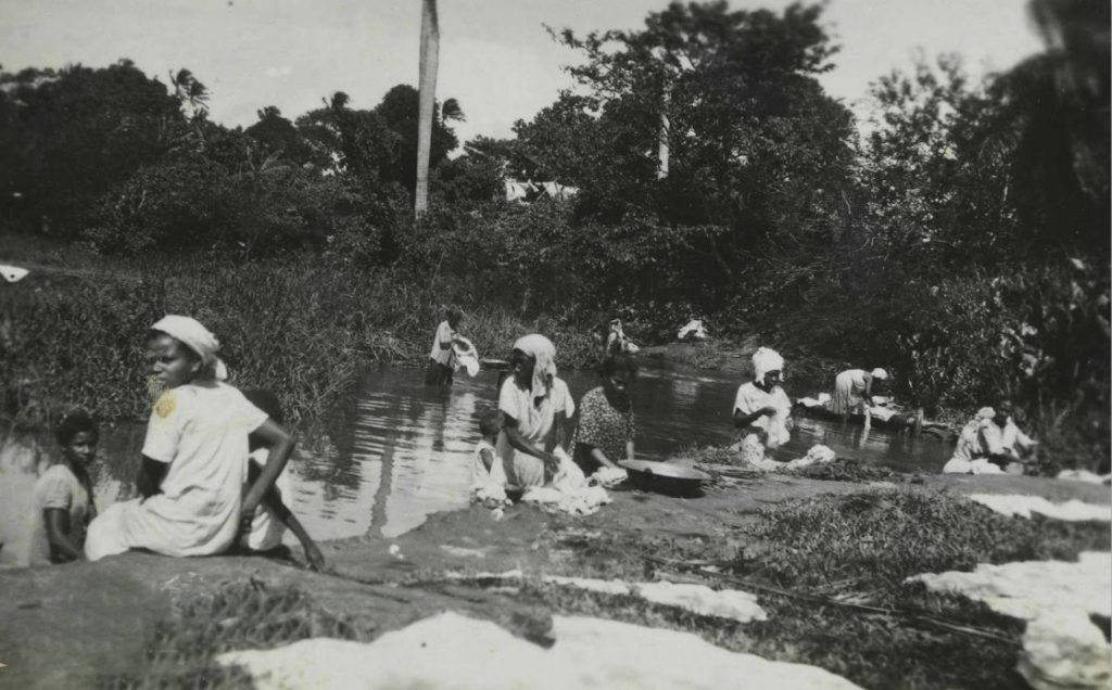 A imagem mostra um grupo de pessoas reunidas às margens de um rio ou riacho, cercado por vegetação densa e árvores altas, incluindo palmeiras. A cena é em preto e branco, o que transmite uma sensação de registro histórico. A maioria das pessoas são mulheres, que estão lavando roupas de forma tradicional: algumas esfregam peças sobre pedras, outras enxaguam diretamente na água, enquanto algumas apenas observam ou descansam próximas. O ambiente é rural e transmite a ideia de uma atividade comunitária, onde o trabalho doméstico se mistura com convivência social em meio à natureza.