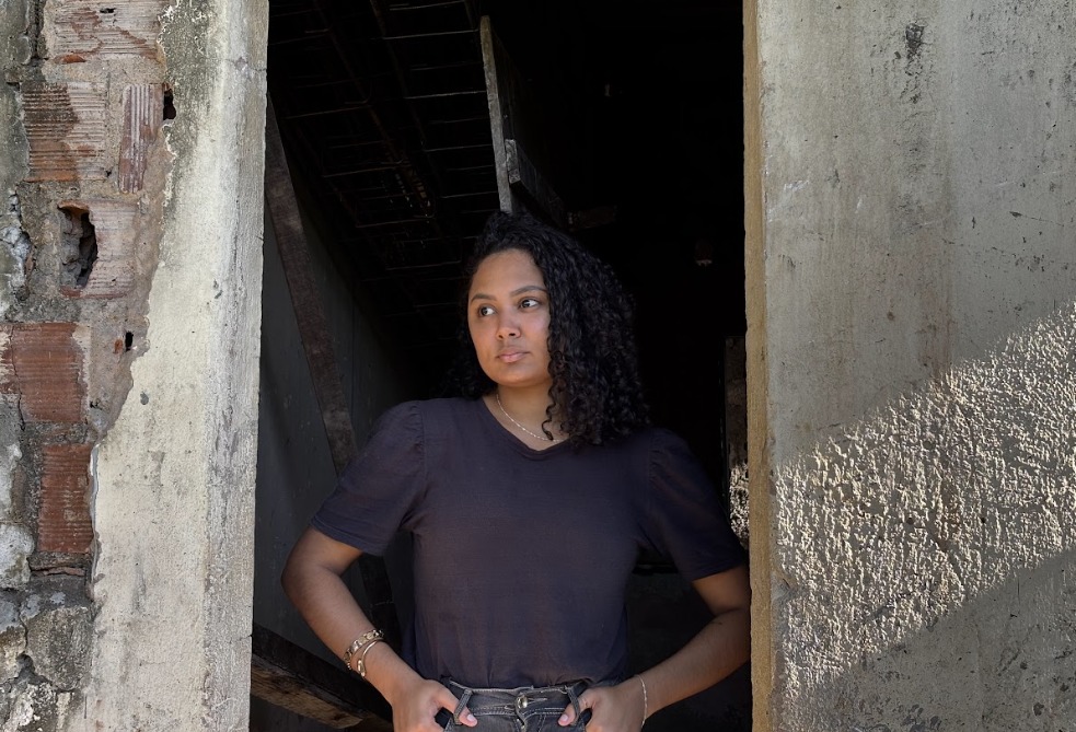 A imagem de Allana Santos mostra uma jovem de cabelos cacheados, vestindo camiseta escura e calça jeans, em pé no vão de uma porta. Ela apoia as mãos no cós da calça, transmitindo uma postura firme e confiante. O prédio onde está é antigo e desgastado: as paredes têm tijolos e concreto expostos, com sinais claros de deterioração. O interior atrás dela é escuro e parece abandonado, com estruturas como vigas e talvez uma escada visíveis na penumbra. O contraste entre a presença da pessoa e o ambiente em ruínas sugere força e resistência.