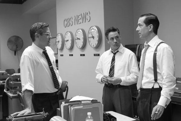 Na cena do filme Boa Noite e Boa Sorte, três homens estão em uma redação de televisão da CBS nos anos 1950, todos vestidos com camisas brancas e gravatas, dois deles usando suspensórios, reunidos em uma conversa séria diante de mesas cheias de máquinas de escrever, pastas e materiais de escritório, enquanto na parede atrás deles há cinco relógios grandes marcando diferentes fusos horários — Londres, Nova York, Chicago, Denver e Los Angeles — reforçando o ambiente de jornalismo intenso e global; a imagem em preto e branco transmite a atmosfera histórica e o clima de concentração e responsabilidade que caracterizam o trabalho dos jornalistas durante o período do macartismo nos Estados Unidos.