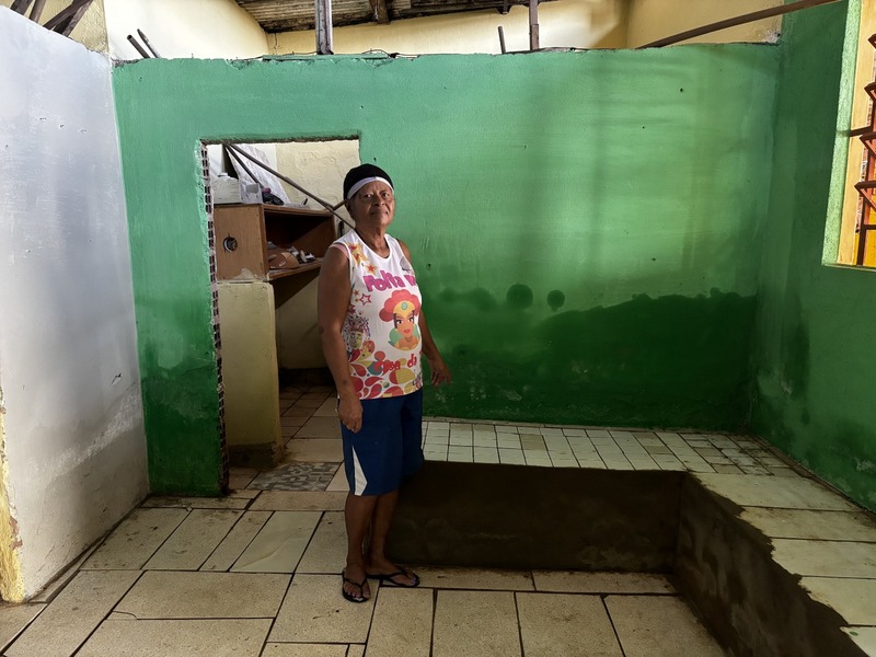 A foto de Laudiceia Silva mostra o interior de um cômodo em reforma, com paredes pintadas de verde e piso de azulejos; no centro há um espaço retangular aberto no chão, como um buraco em obra, e ao fundo vê-se uma porta que leva a outro ambiente com prateleiras e objetos guardados; o teto é de vigas de madeira parcialmente expostas e, à direita, uma janela com grades deixa entrar a luz natural; Laudiceia está em pé, usando uma camiseta colorida com um personagem estampado, além de bermuda azul e sandálias, compondo uma cena que transmite simplicidade e o esforço de melhorar a casa.