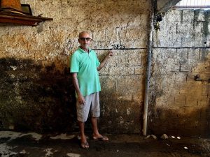 Nivaldo Belmiro aparece em pé dentro de um ambiente fechado, diante de uma parede de blocos de concreto marcada por várias linhas horizontais pretas que registram diferentes alturas da água em enchentes passadas; ele veste camisa polo verde-clara, bermuda bege, chinelos e óculos escuros, e aponta com o dedo para uma dessas marcas, destacando números escritos acima como “15 5.25 15.25” e “14.12”, enquanto a parede mostra manchas e sinais de desgaste, transmitindo a ideia de um registro histórico das cheias que atingiram aquele local.
