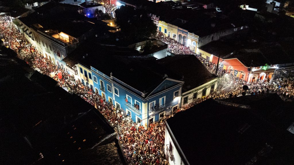 A imagem mostra uma rua de Olinda muito movimentada durante a noite, tomada por uma multidão de pessoas que parecem estar participando de uma grande festa ou celebração. As pessoas estão vestidas com roupas coloridas e ocupam todo o espaço da rua. Os prédios ao redor são pintados em cores vivas como azul, amarelo e vermelho, e a iluminação das construções e dos postes deixa o ambiente brilhante e cheio de energia.