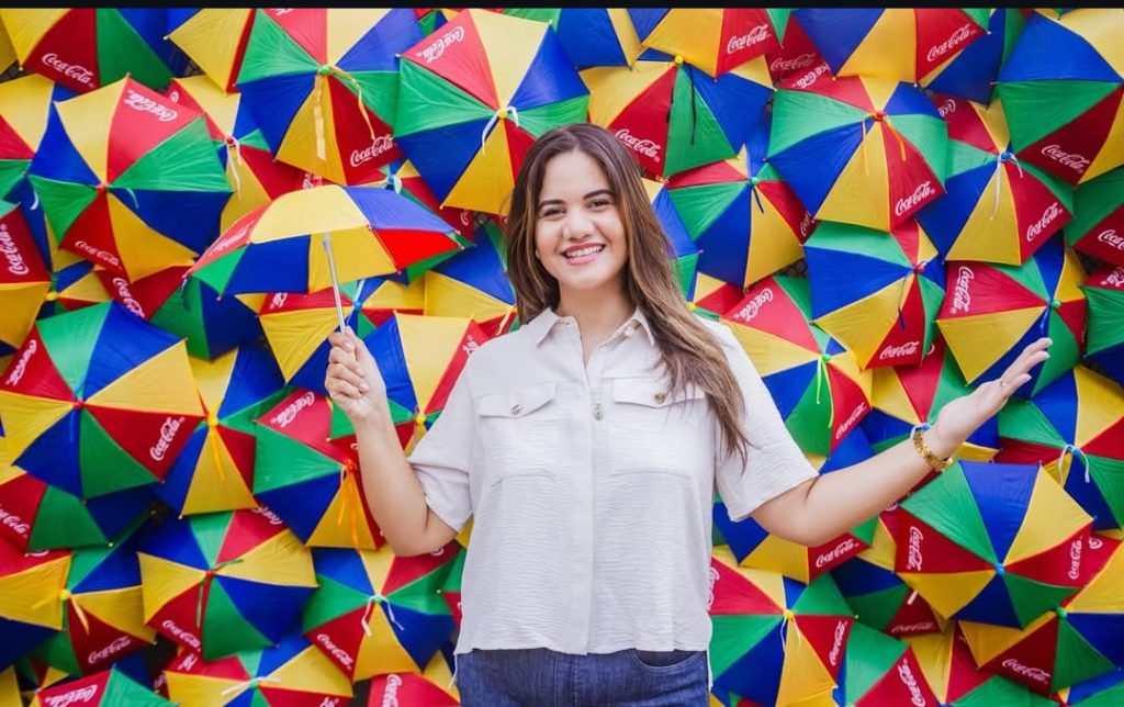 Na foto, vemos a prefeita de Olinda, Mirella Almeida, em frente a um painel vibrante formado por dezenas de pequenos guarda-chuvas coloridos. As peças estão dispostas lado a lado, criando um mosaico alegre de vermelho, amarelo, verde e azul, com muitos dos segmentos vermelhos estampados pelo logotipo da Coca-Cola. Mirella aparece em primeiro plano, vestindo camisa branca e calça jeans azul, segurando um guarda-chuva que combina com o fundo. A cena transmite leveza e descontração, destacando o contraste entre a simplicidade da figura humana e a exuberância cromática da parede. O conjunto cria um ambiente festivo e criativo, típico da atmosfera cultural de Olinda. A prefeita parece integrada ao cenário, como se fosse parte da instalação artística. O guarda-chuva em suas mãos reforça a ideia de continuidade visual com o painel.
