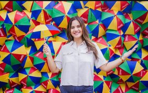 Na foto, vemos a prefeita de Olinda, Mirella Almeida, em frente a um painel vibrante formado por dezenas de pequenos guarda-chuvas coloridos. As peças estão dispostas lado a lado, criando um mosaico alegre de vermelho, amarelo, verde e azul, com muitos dos segmentos vermelhos estampados pelo logotipo da Coca-Cola. Mirella aparece em primeiro plano, vestindo camisa branca e calça jeans azul, segurando um guarda-chuva que combina com o fundo. A cena transmite leveza e descontração, destacando o contraste entre a simplicidade da figura humana e a exuberância cromática da parede. O conjunto cria um ambiente festivo e criativo, típico da atmosfera cultural de Olinda. A prefeita parece integrada ao cenário, como se fosse parte da instalação artística. O guarda-chuva em suas mãos reforça a ideia de continuidade visual com o painel.
