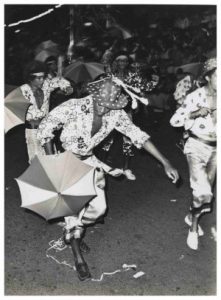 A cena em preto e branco mostra um grupo de pessoas participando de um desfile de carnaval na rua. Elas estão vestidas com roupas estampadas e alegres, e carregam sombrinhas decorativos. Uma pessoa em destaque aparece em movimento, inclinada para frente, usando um chapéu com bolinhas e uma camisa florida, segurando uma sombrinha de frevo. Ao fundo, há mais participantes também com pequenosa guarda-chuvas e roupas vibrantes.