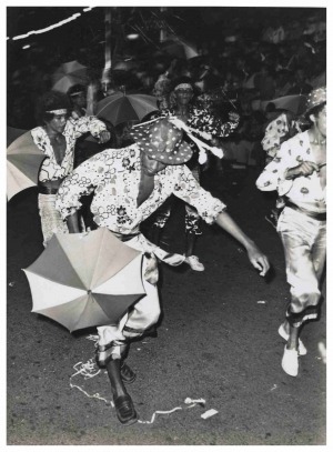 A cena em preto e branco mostra um grupo de pessoas participando de um desfile de carnaval na rua. Elas estão vestidas com roupas estampadas e alegres, e carregam sombrinhas decorativos. Uma pessoa em destaque aparece em movimento, inclinada para frente, usando um chapéu com bolinhas e uma camisa florida, segurando uma sombrinha de frevo. Ao fundo, há mais participantes também com pequenosa guarda-chuvas e roupas vibrantes.